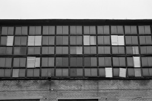 A large industrial building facade with numerous rectangular window panes. Some windows are patched with different materials, indicating wear or repairs. The structure is made of brick, and the general vibe is aged or industrial.