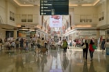A spacious indoor shopping mall area with a high ceiling and illuminated by natural light. People are walking, sitting, and shopping in various stores. There are tables and chairs in the center of the walkway, possibly for dining or resting. Above the central path, a large sign in the middle provides directory information for stores including Myer, David Jones, and Kmart. Electronic advertisements are displayed on large, prominent screens towards the back.