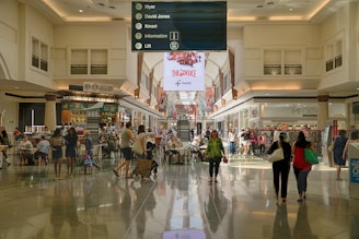 A spacious indoor shopping mall area with a high ceiling and illuminated by natural light. People are walking, sitting, and shopping in various stores. There are tables and chairs in the center of the walkway, possibly for dining or resting. Above the central path, a large sign in the middle provides directory information for stores including Myer, David Jones, and Kmart. Electronic advertisements are displayed on large, prominent screens towards the back.