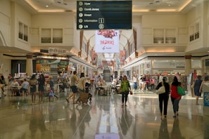 A spacious indoor shopping mall area with a high ceiling and illuminated by natural light. People are walking, sitting, and shopping in various stores. There are tables and chairs in the center of the walkway, possibly for dining or resting. Above the central path, a large sign in the middle provides directory information for stores including Myer, David Jones, and Kmart. Electronic advertisements are displayed on large, prominent screens towards the back.