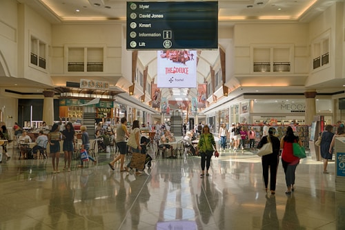 A spacious indoor shopping mall area with a high ceiling and illuminated by natural light. People are walking, sitting, and shopping in various stores. There are tables and chairs in the center of the walkway, possibly for dining or resting. Above the central path, a large sign in the middle provides directory information for stores including Myer, David Jones, and Kmart. Electronic advertisements are displayed on large, prominent screens towards the back.