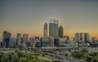 A city skyline with modern skyscrapers standing against the backdrop of a gradient sky transitioning from orange to blue, indicating sunset or sunrise. In the foreground, well-maintained greenery and road infrastructure can be seen. The buildings have varied architectural designs, with glass windows reflecting light.