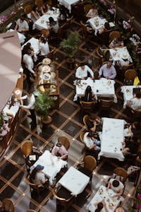 A bustling restaurant setting from an overhead perspective, showcasing several groups of people dining at tables covered with white tablecloths. The space is elegantly decorated with potted plants and floral arrangements lining the sides. Waitstaff can be seen attending to patrons, and a display of pastries is prominently featured on a cart.