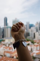A hand wearing a charm bracelet with heart-shaped pendants is raised against a backdrop of a city skyline in daylight.