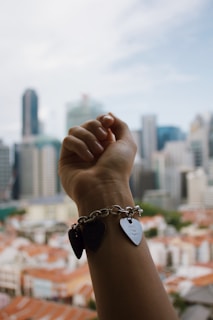 A hand wearing a charm bracelet with heart-shaped pendants is raised against a backdrop of a city skyline in daylight.