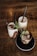 Three beverages are arranged on a wooden table, each placed in a clear glass with creamy liquid and chocolate swirls. The drinks are topped with frothed milk and a sprinkle of cocoa. Fresh mint leaves garnish each glass, and black straws are inserted into the two taller drinks. The shorter drink is served on a black saucer.