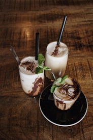 Three beverages are arranged on a wooden table, each placed in a clear glass with creamy liquid and chocolate swirls. The drinks are topped with frothed milk and a sprinkle of cocoa. Fresh mint leaves garnish each glass, and black straws are inserted into the two taller drinks. The shorter drink is served on a black saucer.