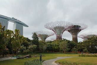 Large futuristic steel structures resembling trees rise above lush greenery. A modern, uniquely designed building is visible in the background, partially covered by plants.
