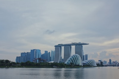 A modern cityscape featuring a prominent skyline with uniquely designed buildings. The Marina Bay Sands complex stands out with its iconic three towers connected by a rooftop platform. Nearby, the futuristic structures of the Gardens by the Bay appear with their domed design. The sky is overcast, and the reflection of the buildings is visible on the calm water in the foreground.