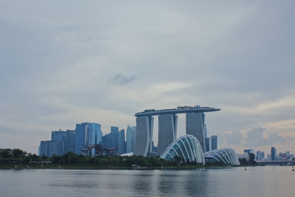 A modern cityscape featuring a prominent skyline with uniquely designed buildings. The Marina Bay Sands complex stands out with its iconic three towers connected by a rooftop platform. Nearby, the futuristic structures of the Gardens by the Bay appear with their domed design. The sky is overcast, and the reflection of the buildings is visible on the calm water in the foreground.