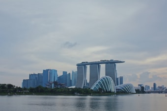 A modern cityscape featuring a prominent skyline with uniquely designed buildings. The Marina Bay Sands complex stands out with its iconic three towers connected by a rooftop platform. Nearby, the futuristic structures of the Gardens by the Bay appear with their domed design. The sky is overcast, and the reflection of the buildings is visible on the calm water in the foreground.