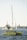 A bright yellow catamaran sails on the sea with a tugboat following in the background. The catamaran has a mast and several people on board. In the distance, a rocky breakwater and a small lighthouse are visible under a clear sky.