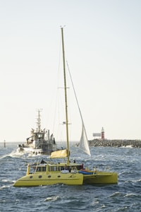 A bright yellow catamaran sails on the sea with a tugboat following in the background. The catamaran has a mast and several people on board. In the distance, a rocky breakwater and a small lighthouse are visible under a clear sky.