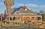 A large, traditional house with a wrap-around veranda and a striking red brick facade is set in a slightly overgrown garden. The veranda features an ornate railing and several arches, creating a picturesque and inviting appearance. A tall, slender palm tree stands prominently in the front, adding a tropical element to the scene. The roof is made of corrugated metal with gabled sections, and a white chimney rises from its center. The sky is clear with a few wispy clouds, suggesting a sunny, tranquil day.