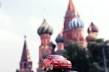 A miniature red BMW X6 toy car is prominently displayed in the foreground with the iconic, colorful domes of St. Basil's Cathedral in Red Square, Moscow, blurred in the background. The clear, bright sky complements the vivid colors of the scene.