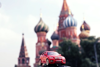 A miniature red BMW X6 toy car is prominently displayed in the foreground with the iconic, colorful domes of St. Basil's Cathedral in Red Square, Moscow, blurred in the background. The clear, bright sky complements the vivid colors of the scene.