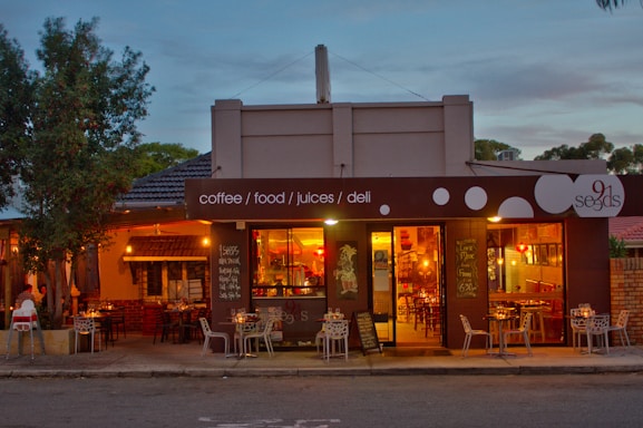 A cozy cafe with a warm, inviting atmosphere featuring outdoor and indoor seating. The exterior has a modern design with large windows allowing a view inside. There are visible signs for coffee, food, juices, and deli offerings next to decorative chairs and tables.