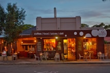 A cozy cafe with a warm, inviting atmosphere featuring outdoor and indoor seating. The exterior has a modern design with large windows allowing a view inside. There are visible signs for coffee, food, juices, and deli offerings next to decorative chairs and tables.