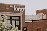 A brick building with large windows and a sign labeled 'the forge' in white letters. The architecture features a classic industrial style. In the background, there are HVAC units on the rooftop. Green leafy trees partially obscure the view, adding a touch of nature to the urban setting.