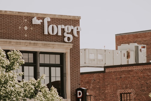 A brick building with large windows and a sign labeled 'the forge' in white letters. The architecture features a classic industrial style. In the background, there are HVAC units on the rooftop. Green leafy trees partially obscure the view, adding a touch of nature to the urban setting.