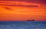 A cargo ship sailing at sunset, silhouetted against vibrant orange and pink clouds.