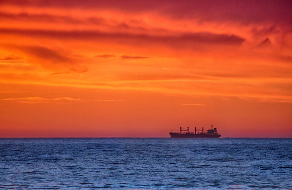 A calm ocean horizon at sunset with a cargo ship silhouetted against the colorful sky.
