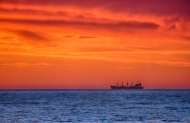 A silhouette of a cargo ship sails on the horizon against a vibrant sunset, where the sky is painted in shades of orange, red, and gold. The calm ocean in the foreground reflects some of the hues from the sky.