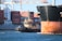 A large cargo ship with containers stacked on deck is accompanied by a tugboat in a harbor. The cargo ship's hull displays visible rust, and cranes are positioned in the background. The water is a vibrant blue, reflecting the sky above.