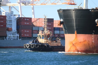 A large cargo ship with containers stacked on deck is accompanied by a tugboat in a harbor. The cargo ship's hull displays visible rust, and cranes are positioned in the background. The water is a vibrant blue, reflecting the sky above.