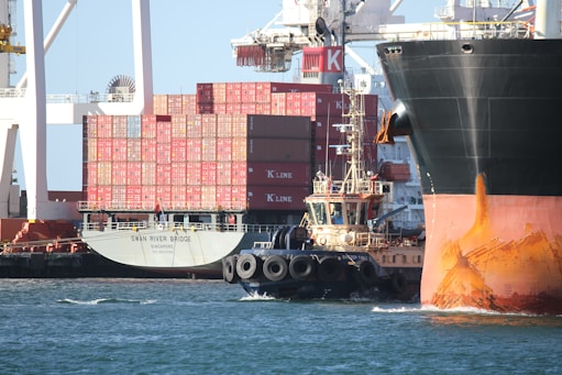 A cargo ship loaded with oil drums navigating through a busy international port.