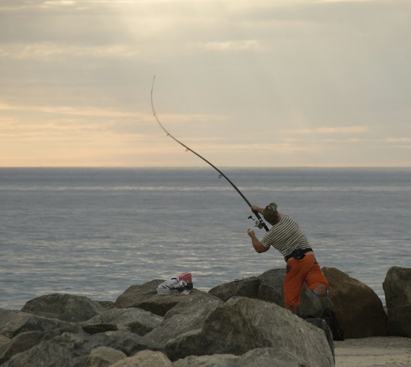 A person is standing on rocks near the shoreline, casting a fishing rod into the open sea. The sky is overcast with a subtle glow from the setting or rising sun, creating a tranquil atmosphere.