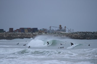 Surfers ride waves near a rocky shoreline, with an industrial backdrop featuring stacked shipping containers and storage tanks. The overcast sky adds a muted tone, and the scene is filled with energy and motion.