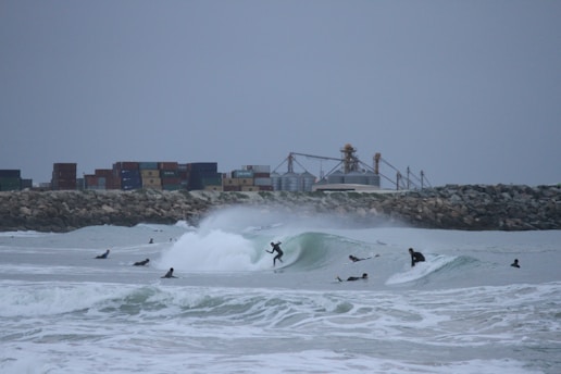 Surfers ride waves near a rocky shoreline, with an industrial backdrop featuring stacked shipping containers and storage tanks. The overcast sky adds a muted tone, and the scene is filled with energy and motion.