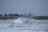 Surfers ride waves near a rocky shoreline, with an industrial backdrop featuring stacked shipping containers and storage tanks. The overcast sky adds a muted tone, and the scene is filled with energy and motion.