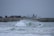 Surfers ride waves near a rocky shoreline, with an industrial backdrop featuring stacked shipping containers and storage tanks. The overcast sky adds a muted tone, and the scene is filled with energy and motion.