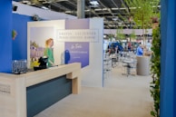 A well-lit indoor exhibition or event space features a counter with a person standing behind it. The decor includes blue walls and potted plants, with a display sign that reads 'Graves, Sauternes, Pessac-Léognan, Barsac.' Several tables and chairs are set up in the background, suggesting a dining or meeting area.