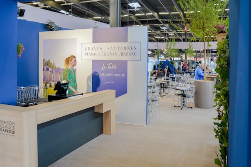 A well-lit indoor exhibition or event space features a counter with a person standing behind it. The decor includes blue walls and potted plants, with a display sign that reads 'Graves, Sauternes, Pessac-Léognan, Barsac.' Several tables and chairs are set up in the background, suggesting a dining or meeting area.