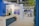 A well-lit indoor exhibition or event space features a counter with a person standing behind it. The decor includes blue walls and potted plants, with a display sign that reads 'Graves, Sauternes, Pessac-Léognan, Barsac.' Several tables and chairs are set up in the background, suggesting a dining or meeting area.
