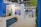 A well-lit indoor exhibition or event space features a counter with a person standing behind it. The decor includes blue walls and potted plants, with a display sign that reads 'Graves, Sauternes, Pessac-Léognan, Barsac.' Several tables and chairs are set up in the background, suggesting a dining or meeting area.