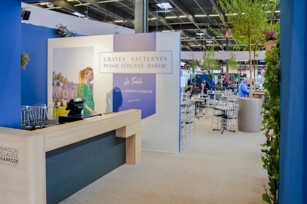 A well-lit indoor exhibition or event space features a counter with a person standing behind it. The decor includes blue walls and potted plants, with a display sign that reads 'Graves, Sauternes, Pessac-Léognan, Barsac.' Several tables and chairs are set up in the background, suggesting a dining or meeting area.