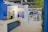 A well-lit indoor exhibition or event space features a counter with a person standing behind it. The decor includes blue walls and potted plants, with a display sign that reads 'Graves, Sauternes, Pessac-Léognan, Barsac.' Several tables and chairs are set up in the background, suggesting a dining or meeting area.