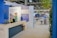 A well-lit indoor exhibition or event space features a counter with a person standing behind it. The decor includes blue walls and potted plants, with a display sign that reads 'Graves, Sauternes, Pessac-Léognan, Barsac.' Several tables and chairs are set up in the background, suggesting a dining or meeting area.