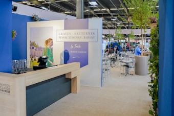 A well-lit indoor exhibition or event space features a counter with a person standing behind it. The decor includes blue walls and potted plants, with a display sign that reads 'Graves, Sauternes, Pessac-Léognan, Barsac.' Several tables and chairs are set up in the background, suggesting a dining or meeting area.