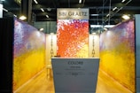 A vibrant exhibition booth features abstract artwork on the walls with a mix of colors such as orange, red, yellow, and purple. The display includes a white counter with text that reads 'Colore Toscana' and 'Bibi Graetz'. Overhead lighting provides ample illumination, and modern ceiling structures add a contemporary vibe.