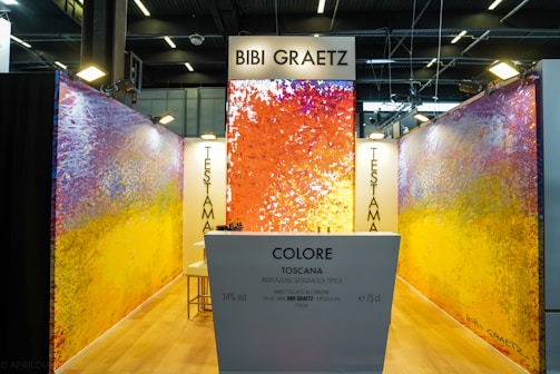 A vibrant exhibition booth features abstract artwork on the walls with a mix of colors such as orange, red, yellow, and purple. The display includes a white counter with text that reads 'Colore Toscana' and 'Bibi Graetz'. Overhead lighting provides ample illumination, and modern ceiling structures add a contemporary vibe.