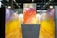 A vibrant exhibition booth features abstract artwork on the walls with a mix of colors such as orange, red, yellow, and purple. The display includes a white counter with text that reads 'Colore Toscana' and 'Bibi Graetz'. Overhead lighting provides ample illumination, and modern ceiling structures add a contemporary vibe.