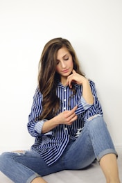woman wearing blue and white striped dress shirt and blue denim jeans sitting on gray surface