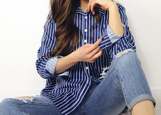 woman wearing blue and white striped dress shirt and blue denim jeans sitting on gray surface