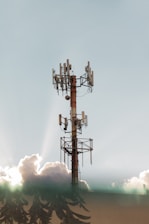 A communications tower stands tall against a clear sky, surrounded by fluffy clouds. The metal structure is equipped with multiple antennas, and the sunlight creates a halo effect through the clouds.