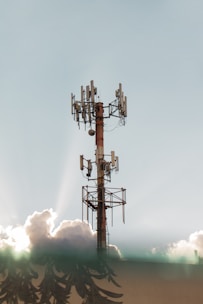 A communications tower stands tall against a clear sky, surrounded by fluffy clouds. The metal structure is equipped with multiple antennas, and the sunlight creates a halo effect through the clouds.
